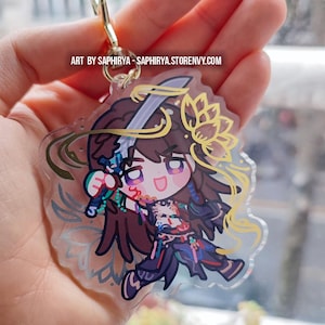 Puede incluir: Un llavero de acr&iacute;lico transparente con un personaje estilo chibi de cabello largo casta&ntilde;o, sosteniendo una espada y vistiendo ropa oscura con detalles coloridos. El dise&ntilde;o incluye una flor de loto amarilla y el texto ART BY SAPHIRYA - SAPHIRYA.STORENVY.COM.