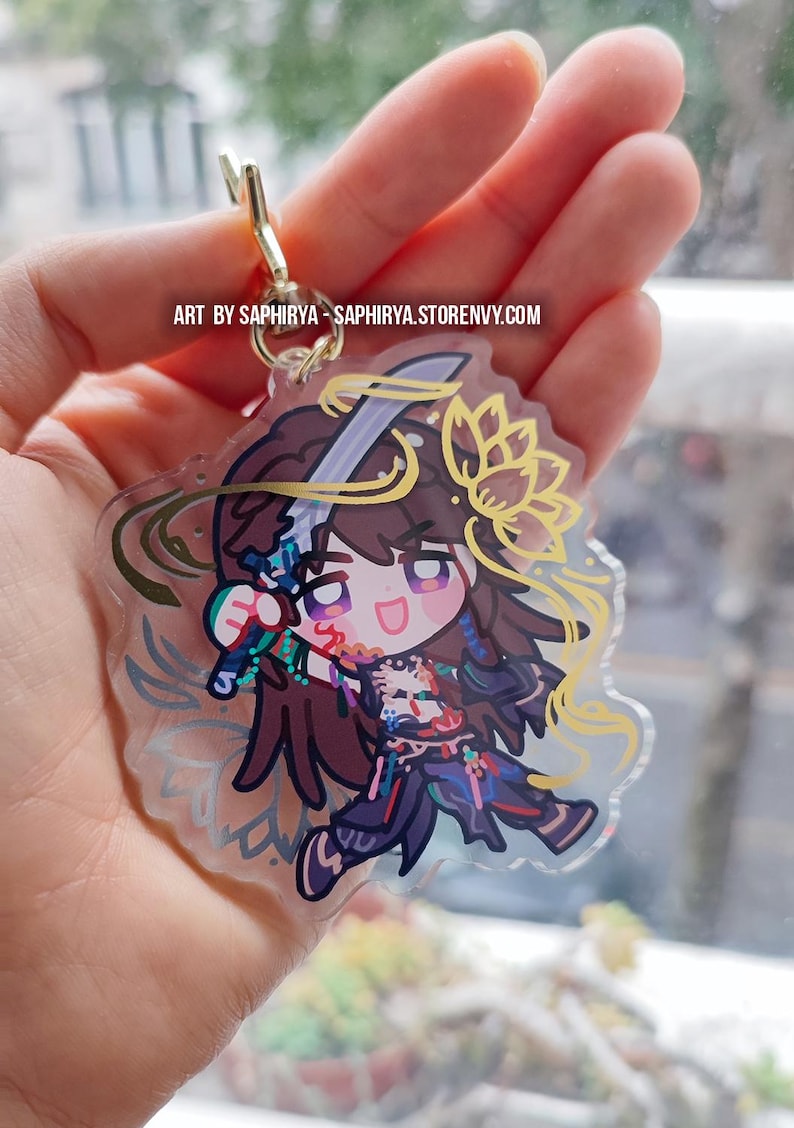 Puede incluir: Un llavero de acr&iacute;lico transparente con un personaje estilo chibi de cabello largo casta&ntilde;o, sosteniendo una espada y vistiendo ropa oscura con detalles coloridos. El dise&ntilde;o incluye una flor de loto amarilla y el texto ART BY SAPHIRYA - SAPHIRYA.STORENVY.COM.