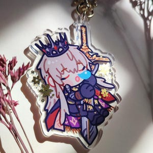 Puede incluir: Un llavero de acr&iacute;lico con un personaje chibi de largo cabello blanco y corona oscura, vistiendo una armadura morada y negra mientras sostiene una gran espada. El dije transparente contiene purpurina dorada en forma de estrella.