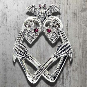 Puede incluir: Colgante plateado con dos calaveras con ojos de gema roja, unidas por un murciélago y manos esqueléticas cruzadas. El colgante tiene detalles intrincados y una estética gótica.