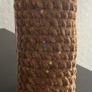 Pode incluir: Um brinquedo de pelúcia marrom crochetado com um rosto sorridente. O brinquedo tem olhos pretos e uma boca preta. O brinquedo é coberto com granulado colorido.