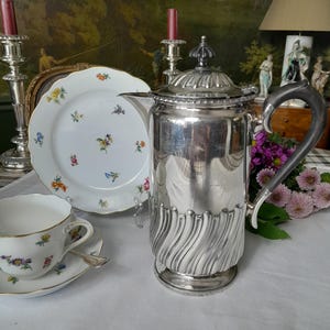 Könnte beinhalten: Eine versilberte Teekanne mit schwarzem Henkel und einem geriffelten Design. Die Teekanne steht auf einer weißen Tischdecke mit Blumenmuster.