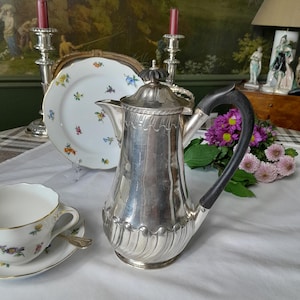 May include: A silver coffee pot with a black handle and an ornate design. The pot is resting on a white tablecloth with a floral pattern.