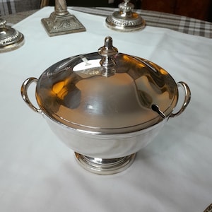 Op de afbeelding: Een zilveren serveerschaal met een deksel en twee handvatten. De schaal heeft een ronde basis en een geribbeld ontwerp. Het deksel heeft een klein tuitje om te gieten.