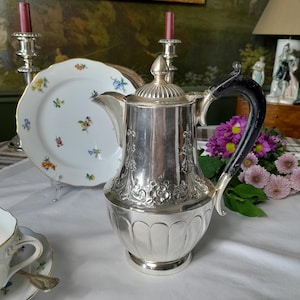 May include: A silver teapot with an ornate design and a black handle. The teapot is sitting on a white tablecloth with a white plate and a cup in the background.