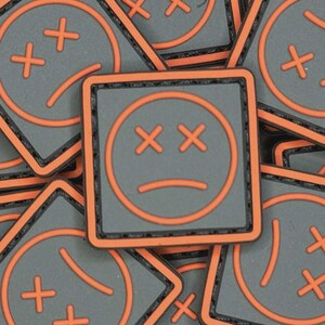 May include: A collection of square patches with a gray background and an orange border. Each patch features a sad face design with crossed-out eyes and a downturned mouth, all in orange. The patches are scattered, creating a textured pattern.
