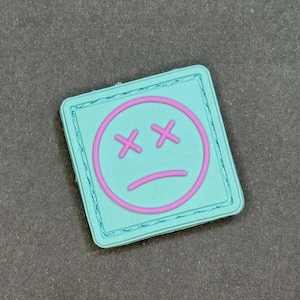 Puede incluir: Parche cuadrado con borde turquesa y diseño de cara triste rosa. La cara presenta ojos tachados y boca hacia abajo. El parche probablemente está hecho de un material duradero.