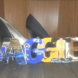 May include: Wooden letters spelling out "MAGEE" in blue, gold, and white, with a pair of black high heels in the background. The letter "A" is decorated with glitter. The letter "G" has the text "Spring 12". The letter "E" has the text "Nothing Just Personal Business".