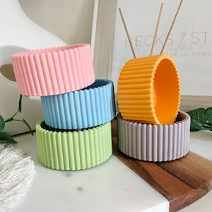 May include: A set of five colourful silicone pot holders in pink, blue, green, grey, and orange. The pot holders have a ribbed texture and are stacked on top of each other.