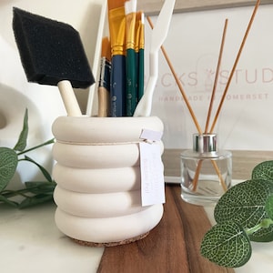 May include: White ceramic bubble pot with a cork base, holding a variety of paint brushes and a sponge brush. The pot has a label that reads "Rocks Studio Bubble Pot".