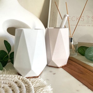 May include: Two geometric concrete planters, one white and one light pink, with cork bottoms. The planters are on a white marble surface with a wooden tray and green foliage.