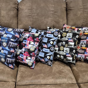 May include: Five square throw pillows featuring various United States military branch themes. The fabric patterns include imagery of Navy ships, Air Force jets, Army tanks, and Marine Corps emblems, all accented with American flags and bald eagles.