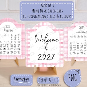 May include: A pack of three mini desk calendars featuring a pink and white plaid border. Two calendars display the January 2027 monthly grid, while the center calendar features the text Welcome to 2027. Each calendar is supported by a small stand.