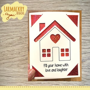 May include: A white card with a red glitter border and a house cutout. The house has a heart in the middle and windows. The text "Fill your home with love and laughter" is printed below the house.