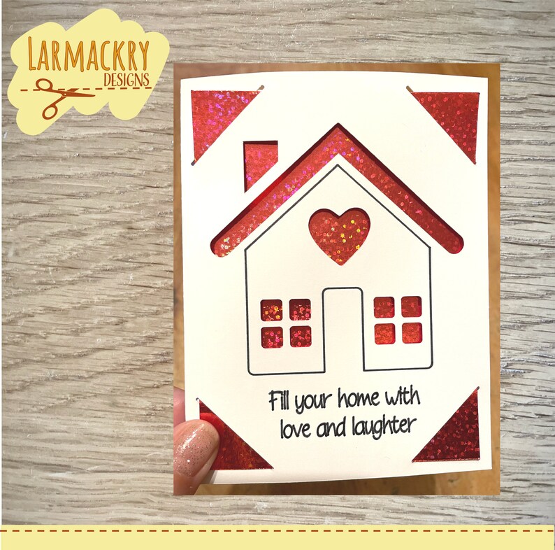 May include: A white card with a red glitter border and a house cutout. The house has a heart in the middle and windows. The text "Fill your home with love and laughter" is printed below the house.