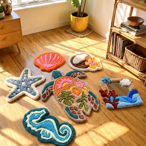 Può includere: Una collezione di sei articoli tessili trapuntati con motivi oceanici, tra cui una conchiglia arancione brillante, una stella marina blu, un cavalluccio marino, una tartaruga marina con motivi floreali, una noce di cocco e uno specchio rettangolare.