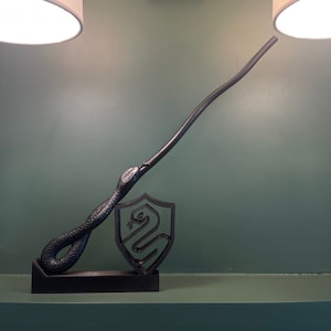 May include: A black wand with a snake design on the handle, resting on a black stand with a snake crest. The wand is curved and pointing upwards. The stand is on a green wall with two gold wall sconces.