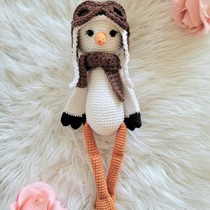 Puede incluir: Muñeca de pájaro de ganchillo hecha a mano con cuerpo blanco, gorro de aviador y bufanda marrones. El pájaro tiene patas y pies naranjas, puntas de ala negras y un pico color melocotón. La muñeca se exhibe sobre una superficie blanca y esponjosa con rosas rosas.
