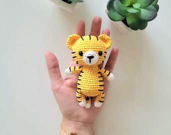 May include: A small, yellow crocheted tiger with black stripes, white paws, and a white muzzle. The handmade tiger is held in a hand, with a gold bracelet with a heart charm visible. Two small potted succulents are in the background.