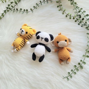 Puede incluir: Tres animales de peluche tejidos a crochet: un tigre rayado amarillo y negro, un panda blanco y negro y un mono marrón. Los animales están sentados sobre una superficie blanca y esponjosa.