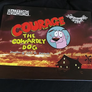 Puede incluir: Portada del libro de la serie animada "Coraje, el perro cobarde". Presenta el título del programa en rojo y amarillo, un personaje de perro rosa y una silueta de casa y molino de viento contra un atardecer.