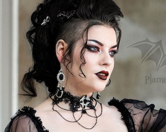 May include: A woman in a black lace and tulle dress with a sheer bodice and long sleeves. She is wearing a silver belt with a bat design and a silver necklace with a cross pendant. The dress has a full skirt and the woman is standing in a doorway. The text "Plamendura Art" is visible in the image.