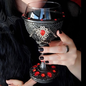 May include: A decorative goblet featuring a textured black finish, ornate silver-toned filigree accents, and multiple embedded red teardrop-shaped gems. The chalice has a wide bowl, a slender stem, and a circular base, all adorned with red stones.