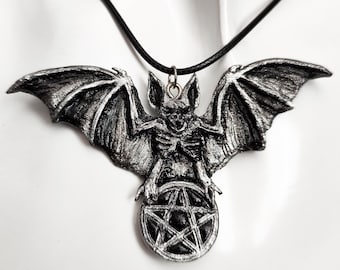 May include: A silver bat pendant with a pentagram charm hanging from a black cord necklace. The bat is depicted with outstretched wings and a skeletal body.