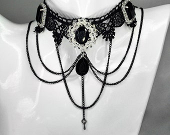 May include: Black lace choker necklace with large black gemstones and dangling chains. The necklace features a central oval gem surrounded by silver-toned embellishments and a teardrop-shaped gem pendant. The brand name "Flamendura Art" is visible.