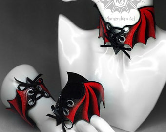 May include: A set of three black and red leather accessories with bat wing designs. The set includes a choker, a bracelet, and a ring. The accessories are laced up the front and feature silver eyelets. The text "Plamendura Art" is visible on the choker.