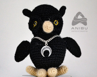 May include: Black crocheted owl with yellow eyes and a silver crescent moon necklace. The owl is sitting on a white surface. Anibu Arts & Crafts is printed on the background.