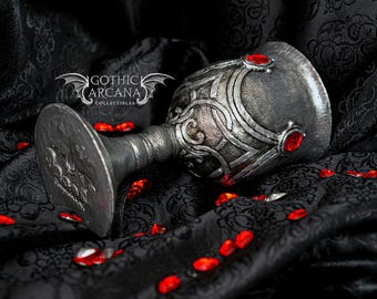 May include: A silver and black goblet with a decorative design and red gemstone accents. The goblet is resting on a black fabric background.