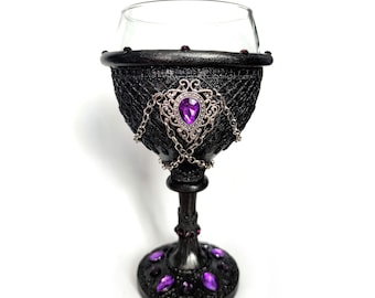 May include: A black glass goblet with a decorative silver and purple design. The goblet has a wide bowl and a tall stem. The bowl is decorated with a silver chain and a purple gemstone. The stem is decorated with a silver chain and purple gemstones.