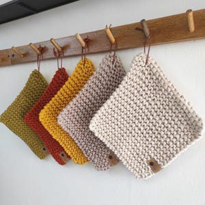 Könnte beinhalten: Sechs gestrickte Topflappen in verschiedenen Farben, darunter Olivgrün, Rost, Gelb, Mauve und Creme. Jeder quadratische Topflappen hängt an einem Holzpflock mit einem Lederetikett. Die Topflappen werden vor einer weißen Wand präsentiert.