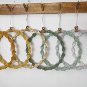 May include: Six macrame hoops in various shades of green and yellow hanging on a wooden rack. The hoops are made of twisted rope and have a natural, bohemian style.