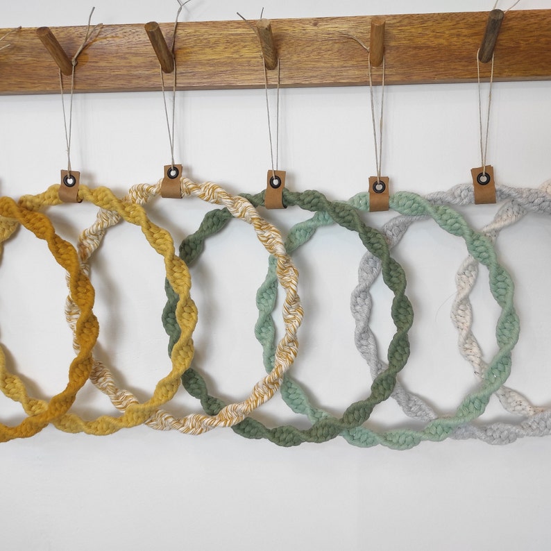 May include: Six macrame hoops in various shades of green and yellow hanging on a wooden rack. The hoops are made of twisted rope and have a natural, bohemian style.