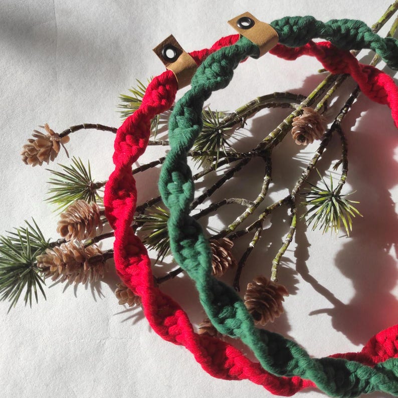 May include: Two braided rope wreaths, one red and one green, are displayed on a branch of a pine tree. The wreaths are made of thick, twisted rope and have a simple, rustic look.