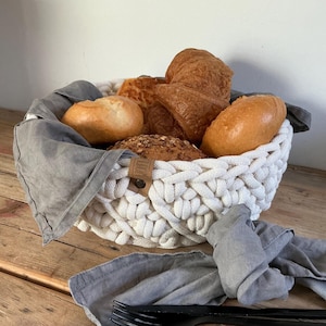 May include: A round, off-white, thick-corded crochet basket lined with a grey fabric cloth, filled with various golden-brown baked bread rolls and croissants, featuring a small tan leather tag with the text 'LUNHOF' on the side.