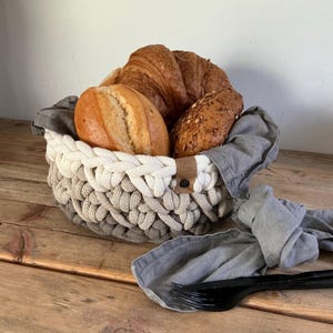 May include: A round, hand-crocheted basket featuring a gradient of white, beige, and gray yarn. The basket is lined with a gray fabric cloth and contains several baked bread rolls and a croissant. A small brown leather tag is attached to the side.