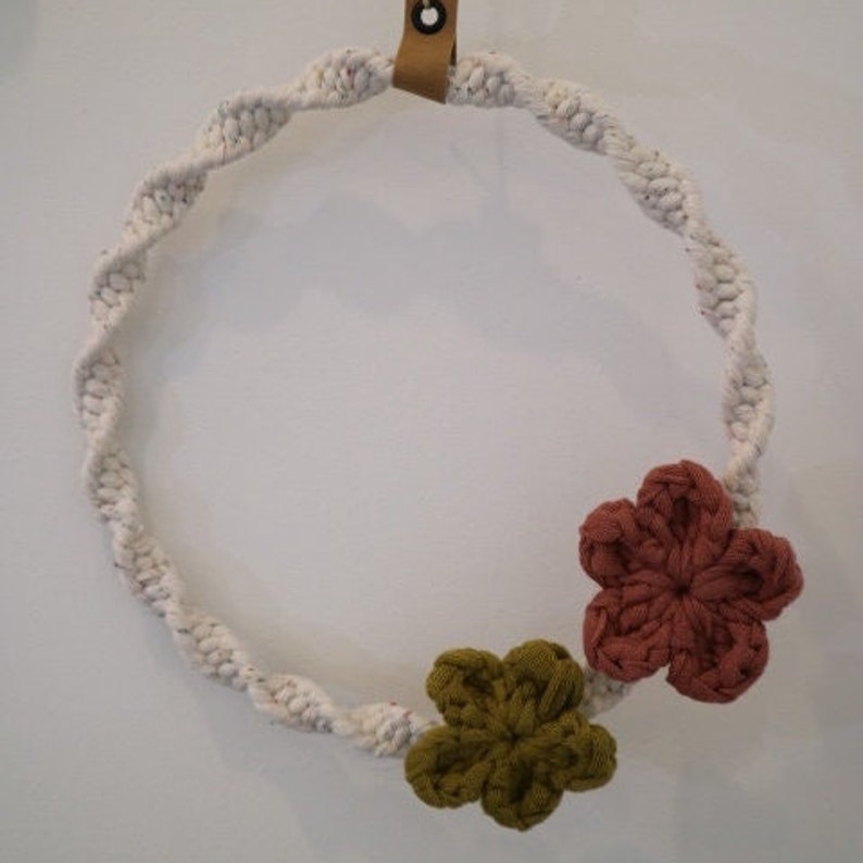 May include: A white rope wreath with two crocheted flowers, one brown and one green, attached to the bottom.