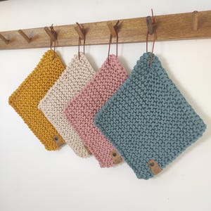 Könnte beinhalten: Vier gestrickte quadratische Waschlappen in Senfgelb, Creme, Rosa und Türkis hängen an einer Holzstange. Jeder Lappen hat ein Lederetikett und eine Schlaufe zum Aufhängen. Die Waschlappen sind aus dickem, strukturiertem Garn gefertigt.