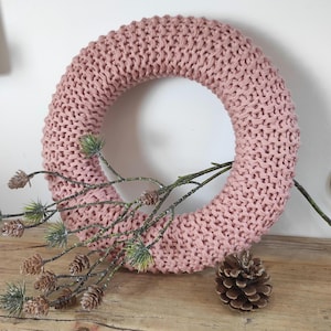 May include: A circular wreath made of chunky, dusty pink knit fabric. The wreath is decorated with a branch featuring small green pine needles and several brown pinecones, with an additional loose pinecone resting near the base of the wreath.