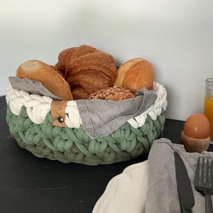 May include: A round, hand-crocheted basket made of thick cotton rope in shades of sage green and cream. The basket contains several golden-brown baked goods, including croissants and a seeded roll, lined with a gray fabric napkin.