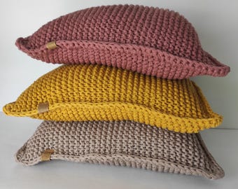 Könnte beinhalten: Ein Stapel aus drei rechteckigen Häkelkissen in Altrosa, Senfgelb und Beige. Jedes Kissen weist ein strukturiertes Strickmuster, einen dicken gehäkelten Rand und ein kleines beiges Etikett in Lederoptik an der Seitenkante des Bezugs auf.