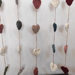 May include: A garland of crocheted hearts in various colors, including white, red, green, and brown, hangs from a white wall. The hearts are strung on twine with small wooden beads.