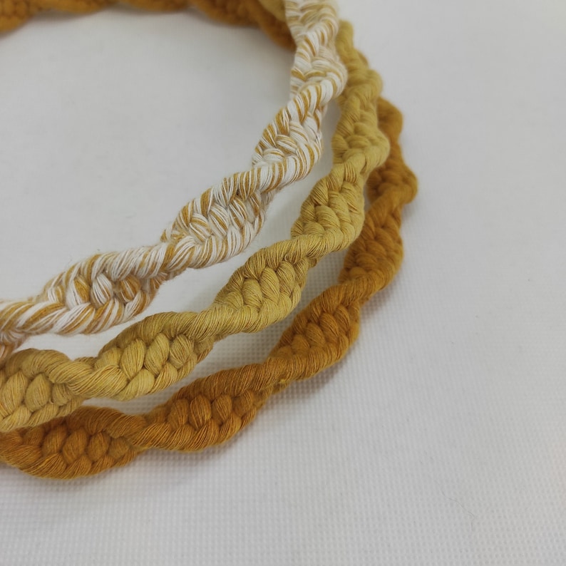 May include: Three braided cords in shades of yellow and white, woven together to form a circular shape. The cords are twisted and knotted, creating a textured and intricate design.