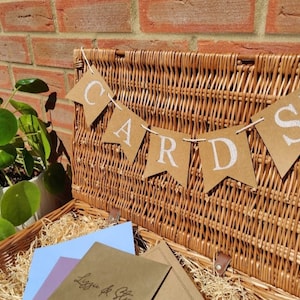 May include: A wicker basket with a brown paper banner that says "CARDS" hanging above it. The basket is filled with brown paper and has a few envelopes inside.