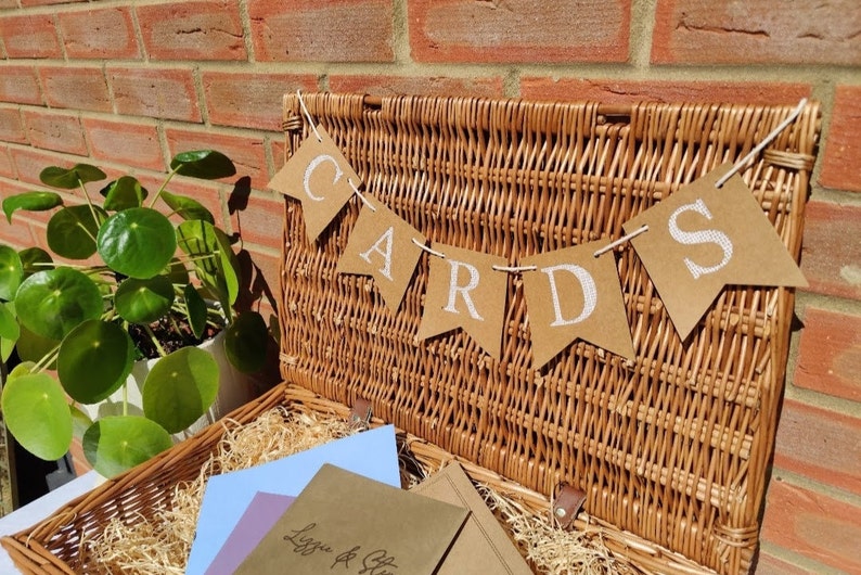 May include: A wicker basket with a brown paper banner that says "CARDS" hanging above it. The basket is filled with brown paper and has a few envelopes inside.