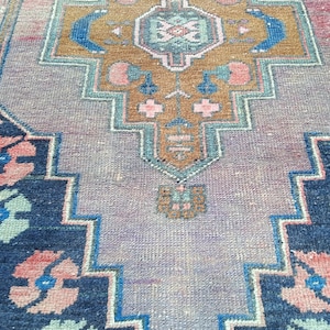 Może przedstawiać: Prostokątny dywan w stylu vintage ze skomplikowanym geometrycznym wzorem. Dywan ma centralny wzór w odcieniach brązu, różu i niebieskiego, otoczony granatową obwódką z motywami kwiatowymi. Ogólna paleta kolorów dywanu jest stonowana.