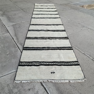 May include: A cream-coloured rug with black horizontal stripes. The rug has a fringed edge and a simple, geometric design. The rug is placed on a stone surface.